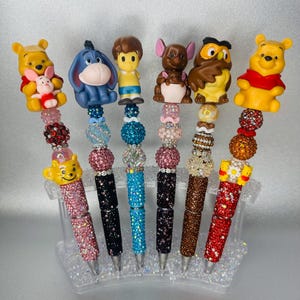 May include: Six pens featuring character toppers including Winnie the Pooh, Eeyore, Christopher Robin, Roo, Owl, and Piglet. Each pen barrel is decorated with colourful, sparkling beads and rhinestones in various textures, patterns, and vibrant hues.