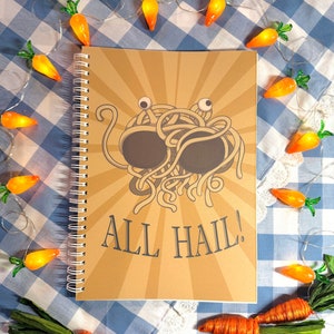 Könnte beinhalten: Ein braunes Spiralnotizbuch mit einem Cartoon-Bild eines Spaghetti-Monsters mit zwei Augen und dem Text "ALL HAIL!" Das Notizbuch ist von einer Lichterkette in Form von Karotten umgeben.