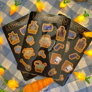 Puede incluir: Tres hojas de pegatinas negras con varios diseños de dibujos animados y el texto "LETHAL COMPANY". Las pegatinas presentan imágenes de criaturas, herramientas y frases como "Yippie!" y "Hugs?". El fondo incluye luces en forma de zanahoria y un patrón de cuadros azules y blancos.