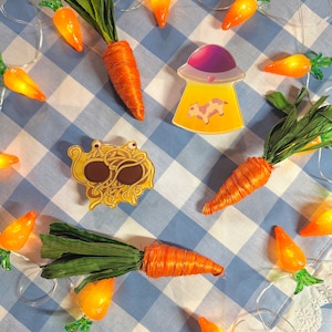May include: A collection of carrot-shaped string lights, a UFO pin with a cow inside, and a spaghetti monster pin. The pins are on a blue and white checkered tablecloth.