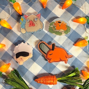 Könnte beinhalten: Fünf bunte Emaille-Pins mit Anime-Charakteren. Die Pins sind auf einem blau-weiß karierten Hintergrund mit einer Lichterkette in Form von Karotten angeordnet.
