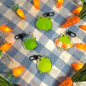 May include: Four green frog keychains with different expressions and accessories. The keychains are hanging from a string of lights with orange carrot-shaped bulbs.
