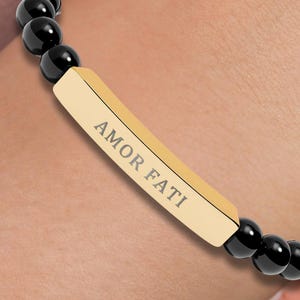 May include: A beaded bracelet featuring black beads and a gold-colored rectangular charm. The charm is inscribed with the words "AMOR FATI". The bracelet is worn on a wrist.