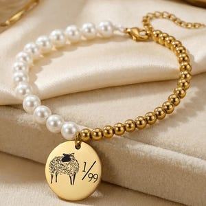 May include: Gold-toned bracelet featuring a combination of white pearls and small gold beads. A circular charm with a sheep illustration and the text "1/99" hangs from the bracelet. The bracelet is displayed on a cream-colored fabric.
