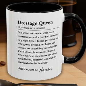 以下が含まれることがあります： 黒い内側とハンドルが付いた白いコーヒーマグ。マグには「Dressage Queen」のテキストと定義、そして「Also known as Karen」という言葉が書かれています。コーヒー豆とスプーンが木製の表面にあります。