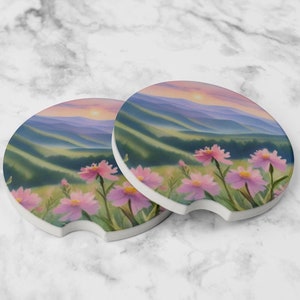 May include: Two round ceramic coasters with a scenic mountain landscape and pink flowers in the foreground. The coasters feature a sunset sky with a soft pink and purple gradient.