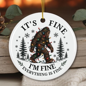 May include: A round white ceramic ornament featuring a brown Bigfoot figure wrapped in colorful Christmas lights, surrounded by black pine trees and snowflakes, with the text IT'S FINE, I'M FINE, EVERYTHING IS FINE printed in black.