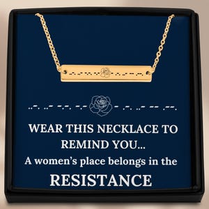 May include: A gold-colored necklace with a bar pendant, presented in a black box with a blue interior. The pendant has a rose design and Morse code. The text reads: "WEAR THIS NECKLACE TO REMIND YOU... A women's place belongs in the RESISTANCE."