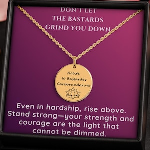 Può includere: Una collana color oro con un ciondolo rotondo. Il ciondolo presenta le parole "Nolite te Bastardes Carborundorum" e un simbolo di fiore di loto. La collana è esposta in una scatola nera con il testo "DON'T LET THE BASTARDS GRIND YOU DOWN."