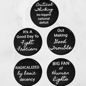 Może przedstawiać: Pięć czarnych okrągłych guzików z białym tekstem. Guziki zawierają hasła takie jak "Critical Thinking the biggest national deficit", "It's a Good Day To Fight Fascism" i "Big Fan of Human Rights".