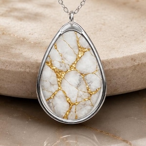 May include: A silver necklace featuring a teardrop-shaped pendant with a white stone inlay accented by gold-colored veining. The pendant hangs from a delicate silver chain with small, evenly spaced metal beads along the length of the chain.