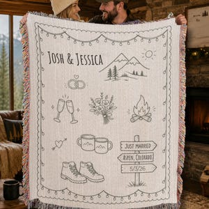 May include: A cream-colored woven throw blanket with a decorative border and fringe. The blanket features a mountain scene, wedding rings, champagne glasses, flowers, a campfire, mugs, hiking boots, and the names "Josh & Jessica". The blanket also has the text "Just Married" and "Aspen, Colorado".