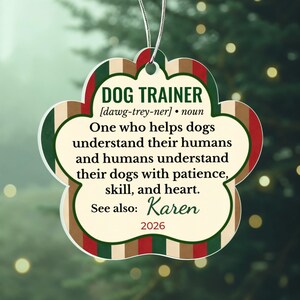 May include: A paw-shaped ornament featuring a striped border in red, green, and tan. The text reads: DOG TRAINER [dawg-trey-ner] noun. One who helps dogs understand their humans and humans understand their dogs with patience, skill, and heart. See also: Karen 2026.