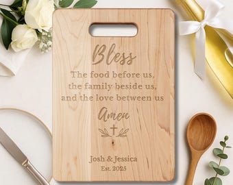 Puede incluir: Tabla de cortar rectangular de madera con asa, grabada con la palabra "Bless" y una oración. Los nombres "Josh & Jessica" y el año "Est. 2025" también están grabados. Se ven una cuchara de madera y un cuchillo.