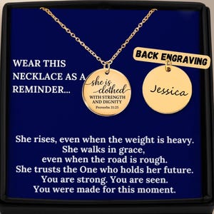 Könnte beinhalten: Goldfarbene Halskette mit zwei runden Anhängern. Ein Anhänger trägt den Text "she is clothed with strength and dignity" und "Proverbs 31:25". Der andere Anhänger hat den Namen "Jessica". Die Halskette wird in einer schwarzen Box mit inspirierendem Text präsentiert.