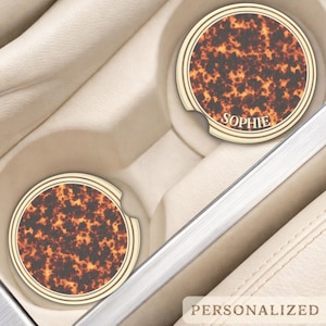 May include: Two personalised car coasters with a tortoiseshell pattern. One coaster has the name "Sophie" printed on it. The coasters are round with a cream-coloured border and are placed in a car's cup holders.
