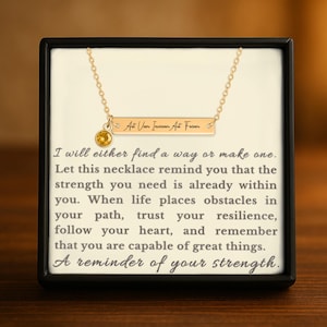 May include: Gold-colored necklace with a rectangular pendant engraved with the phrase "Ast Viam Inveniam Aut Feciam" and a small yellow gemstone. The necklace is displayed in a black box with inspirational text.