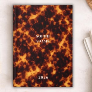 May include: A hardback journal with a tortoiseshell design in brown and yellow hues. The journal displays the name "SOPHIE ADAMS" and the year "2026" in white text. A gold and white pen is positioned on the left.