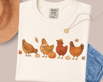 May include: Cream-colored Comfort Colors t-shirt featuring a fall-themed design. The graphic depicts four chickens with pumpkins and autumn leaves. The shirt has a tag that reads "COMFORT COLORS BEST HUGEN M/M". A gold necklace and a brown hat are also visible.