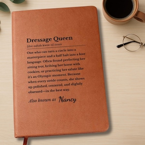 A tan faux leather journal titled "Dressage Queen" is displayed on a light wooden desk. The cover features a humorous and heartfelt definition of a dressage rider, mentioning her love for perfecting the sitting trot, bribing her horse with cookies, & treating her salute like an Olympic event. The phrase "Also known as Nancy" is engraved at the bottom in script. The scene includes a pair of glasses, a pen, a cup of black coffee, & a sprig of eucalyptus, creating a cozy equestrian-themed flat lay.