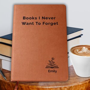 May include: A brown leather journal with the text "Books I Never Want To Forget" and the name "Emily". A pen, books, and a cup of coffee are on a wooden table.