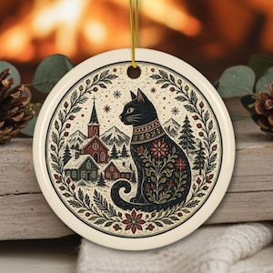 Puede incluir: Un adorno de cerámica redondo con un gato negro con patrones florales sentado en una escena de pueblo invernal. El diseño incluye una iglesia, casas cubiertas de nieve, pinos y montañas, enmarcados por una corona circular de hojas y bayas.