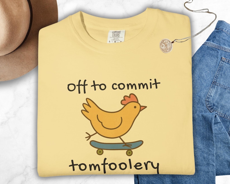 Butter Comfort Colors t-shirt with a quirky chicken riding a skateboard and the phrase ‘off to commit tomfoolery’ in playful handwritten-style text. Funny silly animal design with cottagecore and Gen Z meme vibes.