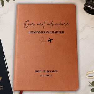 May include: A brown leather journal with the text "Our next adventure HONEYMOON CHAPTER" and the names "Josh & Jessica" with the date "5.9.2025". A heart and airplane graphic are also present. Two passports and wedding rings are visible.