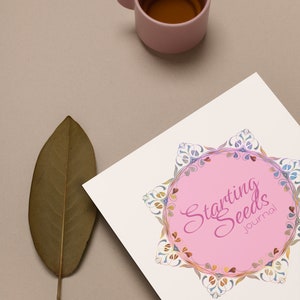 May include: A white journal with a pink circle and the text "Starting Seeds Journal" in pink cursive lettering. The circle is surrounded by a colorful floral design. A brown leaf and a pink cup of tea are in the background.