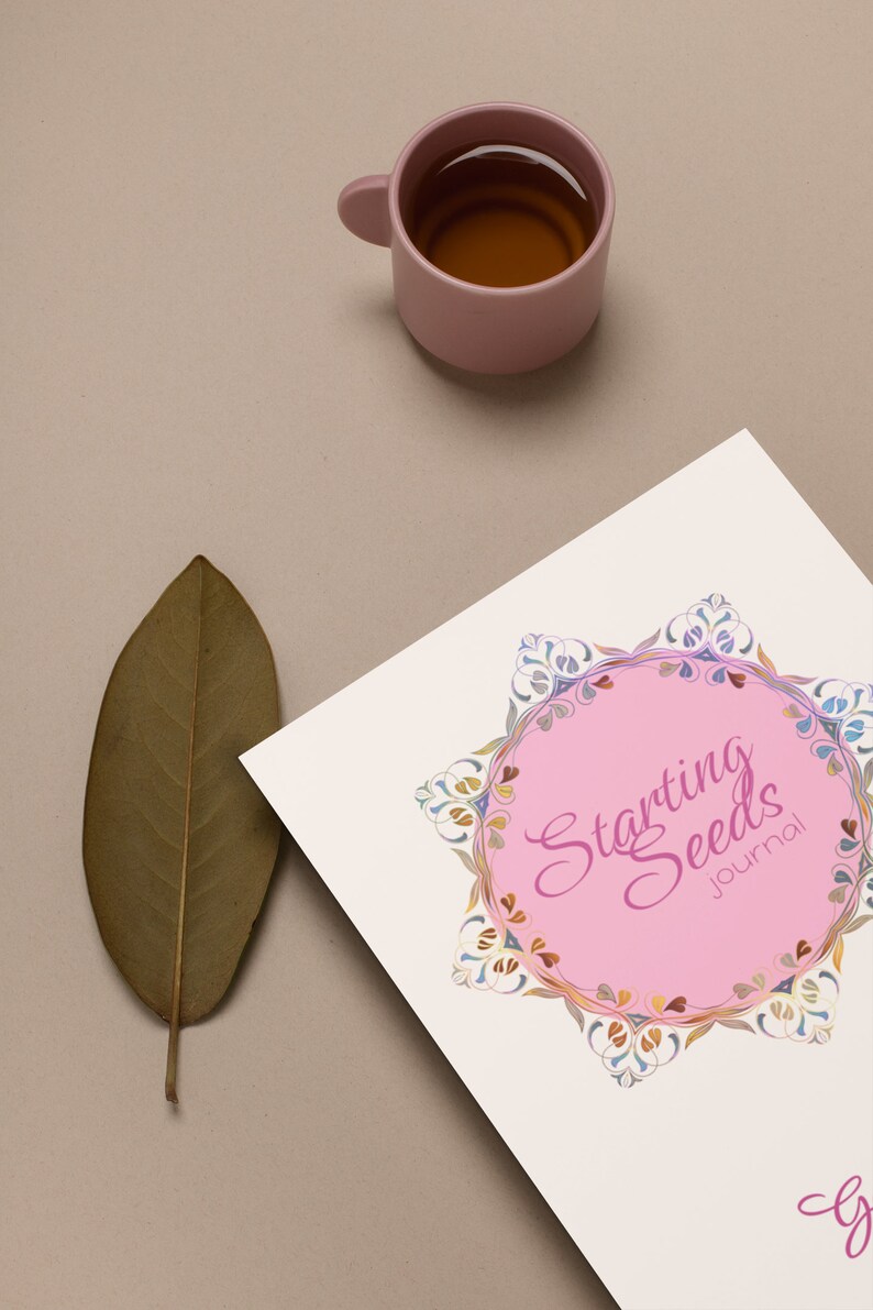 May include: A white journal with a pink circle and the text "Starting Seeds Journal" in pink cursive lettering. The circle is surrounded by a colorful floral design. A brown leaf and a pink cup of tea are in the background.