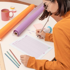 May include: A person is writing on a gridded sheet of paper with a pencil. The paper has the words "Mood Tracker" and "June" printed on it. There are two rolls of wrapping paper, one orange and one purple, a pink sticky note, a ruler, a pink mug, and three colored pencils on the table.