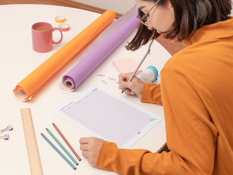 May include: A person is writing on a gridded sheet of paper with a pencil. The paper has the words "Mood Tracker" and "June" printed on it. There are two rolls of wrapping paper, one orange and one purple, a pink sticky note, a ruler, a pink mug, and three colored pencils on the table.