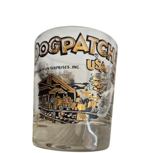May include: Clear glass shot glass with the words "DOGPATCH USA" in gold lettering. The glass features a gold and black illustration of a train and a building. The text "©E CAPP ENTERPRISES, INC." is also visible.