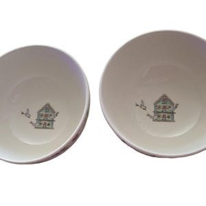May include: Two off-white ceramic bowls. Each bowl features a detailed illustration of a birdhouse with birds in flight. The birdhouse has a green roof and floral accents. The bowls are ideal for serving cereal or soup.