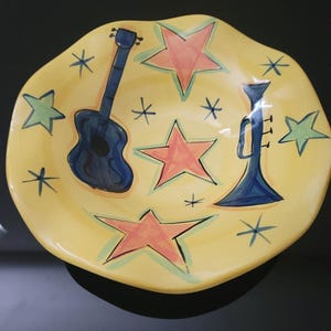 May include: A yellow ceramic serving dish with a wavy edge. The dish is decorated with a blue guitar, a blue trumpet, orange stars, and green stars. Black asterisk shapes are scattered around the design.