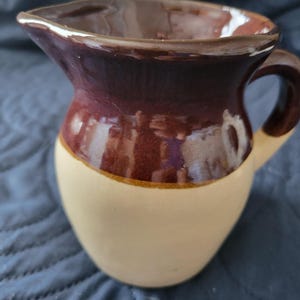 May include: A small, ceramic pitcher with a handle and spout. The pitcher is two-toned, with a cream-colored base and a dark brown upper section. The rim and handle are also dark brown. The pitcher has a glossy finish.