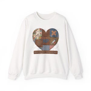 Könnte beinhalten: Ein weißes Sweatshirt mit Rundhalsausschnitt und einem großen, zentralen herzförmigen Motiv, das aus verschiedenen stoffartigen Flicken besteht. Das Design umfasst medizinische Symbole, ein Stethoskop, eine Spritze und die Buchstaben NP, mit dem Text Nurse Practitioner - Healing with Heart auf einem braunen Banner am unteren Rand des Herzens.