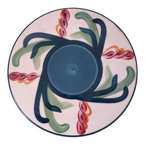 May include: A round, decorative plate with a white background and a dark blue rim. The plate features a central dark blue circle and stylized green and dark blue floral designs, with accents of pink and orange.