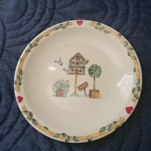 May include: A round, off-white ceramic plate with a decorative birdhouse and garden scene. The rim features a yellow border with green vines and red hearts. The plate's center depicts a birdhouse, trees, and plants.