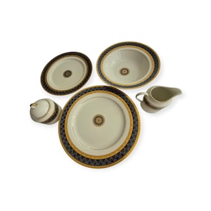 May include: A cream-colored dinnerware set with gold and navy blue geometric patterns. The set includes a large platter, two oval serving dishes, a sugar bowl with a lid, a serving bowl, and a creamer.