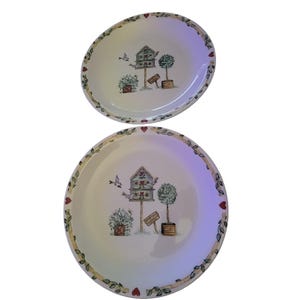 May include: Two white oval plates with a decorative border of leaves and hearts. Each plate features a birdhouse, topiary, and potted plants illustration. The plates are perfect for serving appetizers or desserts.