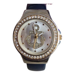 May include: Silver-toned wristwatch with a navy blue band. The watch face features a silver background with a sparkling anchor design. The bezel is encrusted with clear crystals. The watch has gold-colored accents.