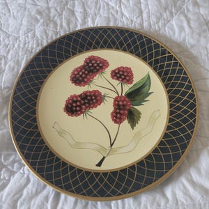 May include: Decorative plate with a cream-colored center featuring a blackberry branch with red berries and green leaves. The rim has a black lattice design with a gold border. A white ribbon is tied around the blackberry stem.