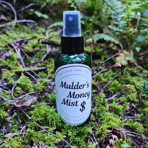 May include: A green glass spray bottle with a black cap and a white label. The label reads "Mulder's Money Mist" with a dollar sign symbol. The words "Prosper and Thrive" and "You Are Sovereign" are also on the label. The bottle is set against a backdrop of green moss.