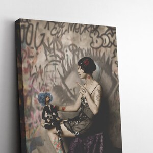 May include: A woman in a black dress and a red flower in her hair sits on a purple cushion. She is holding a doll with blue hair and a black dress. The background is a wall with graffiti.