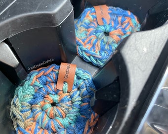 May include: Two crocheted cup coasters in a car cup holder. The coasters are blue, green, and orange with a brown leather tag that says "GraceCreatesCraftCo".