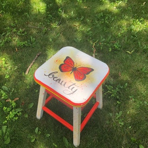 May include: A white wooden stool with red and yellow accents. The top of the stool is painted with a red butterfly and the word "beauty" in black cursive writing.