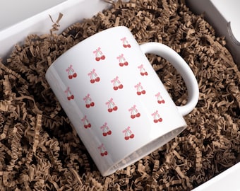 May include: White ceramic mug with a pink and red cherry pattern. The mug is in a white box with brown packing paper.
