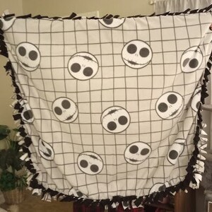 Puede incluir: Una manta de forro polar blanca con un patr&oacute;n de cuadr&iacute;cula blanco y negro e im&aacute;genes repetidas de la cara de Jack Skellington de Pesadilla antes de Navidad. La manta tiene un borde de flecos anudados.