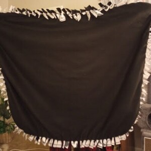 Puede incluir: Una manta de forro polar negra con un borde de flecos anudados blancos. La manta est&aacute; doblada por la mitad y puesta plana sobre una superficie.